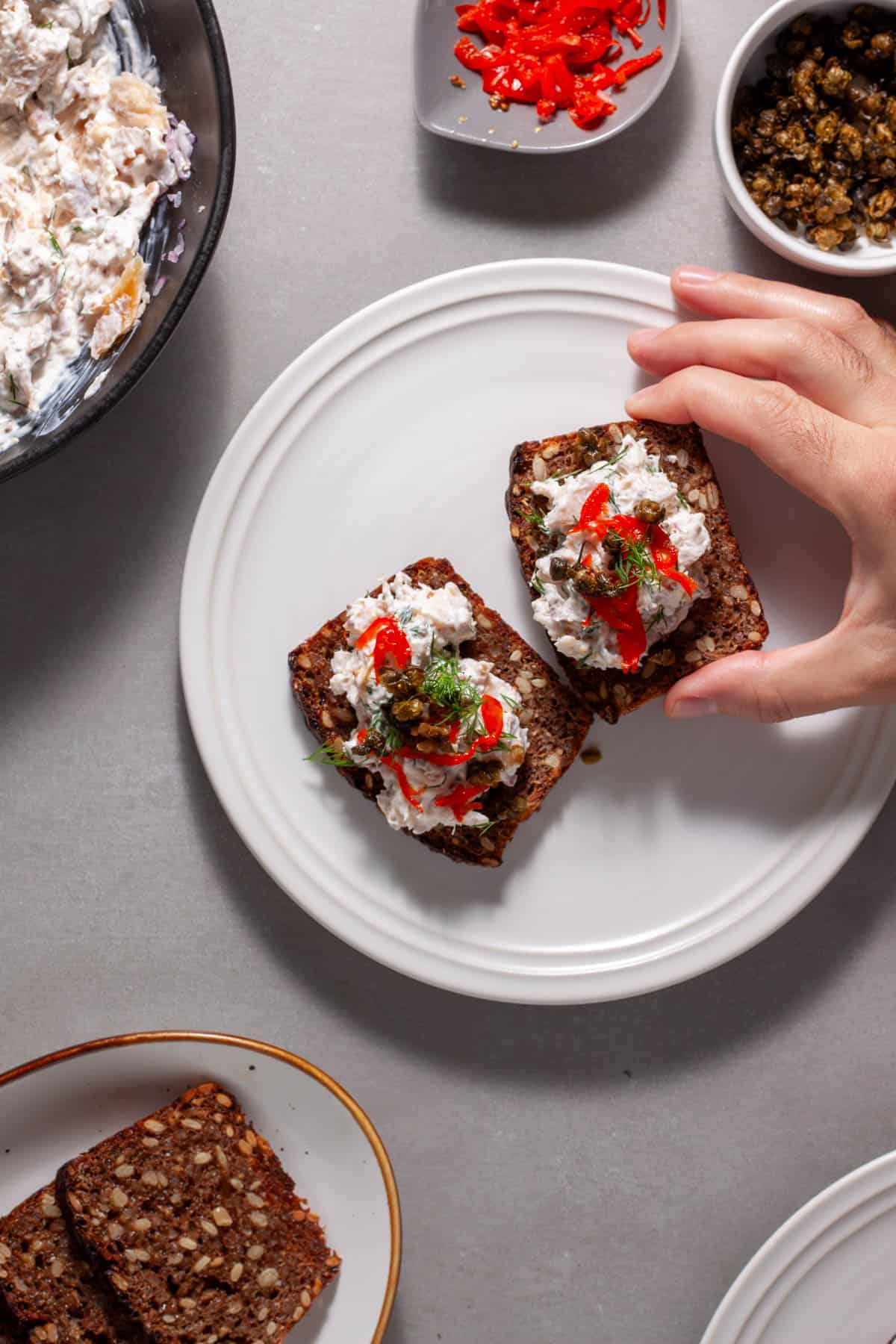 Smoked trout tartine with crispy capers getting assembled on a white plate.