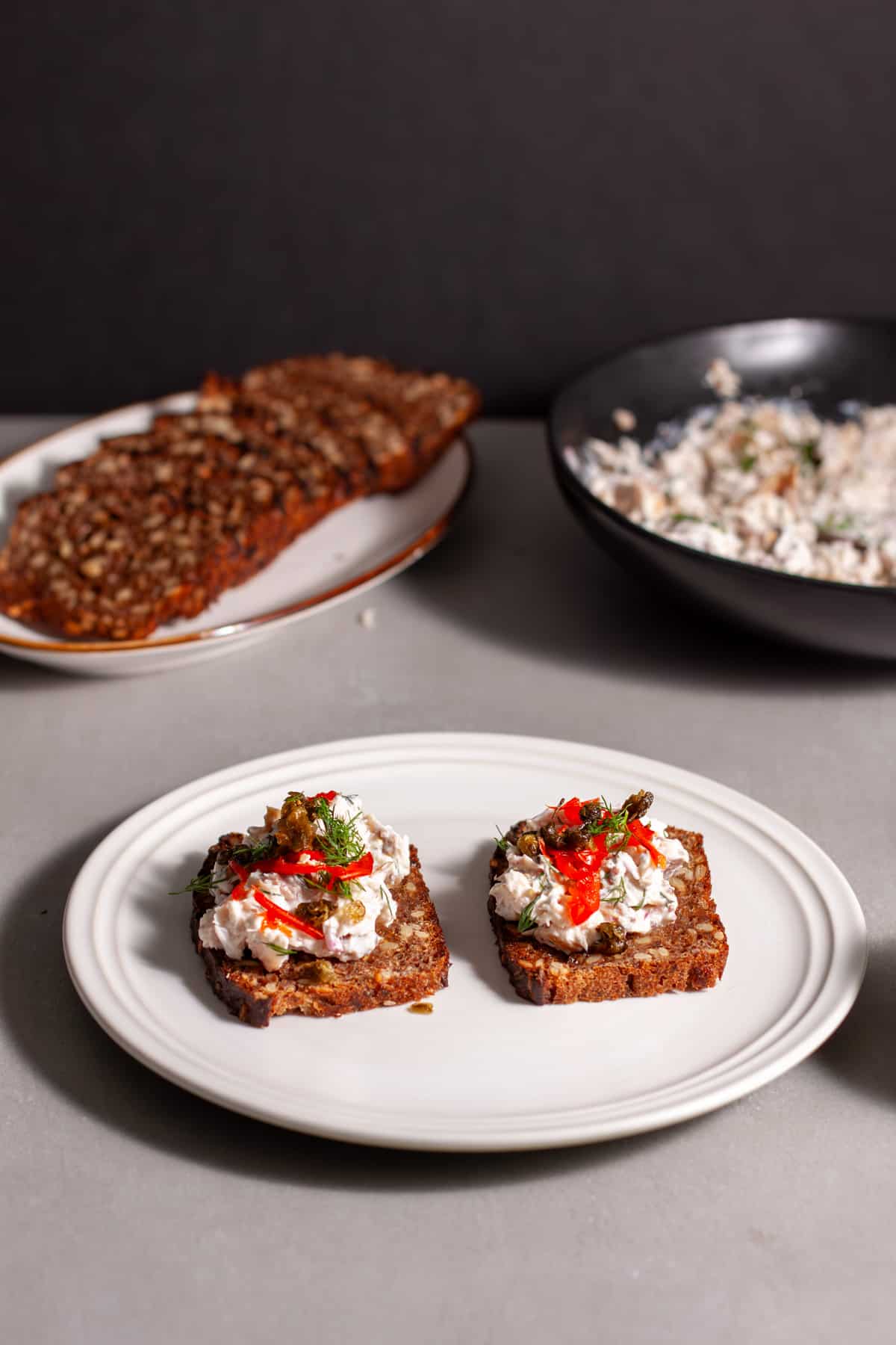 Smoked trout tartines with crispy capers on a small white plate with more toast and fish in the background.