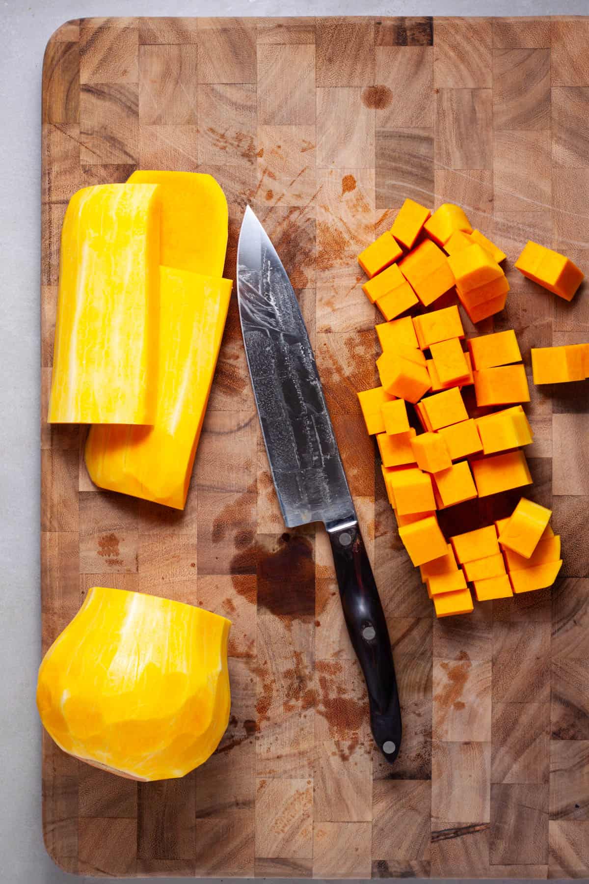 Butternut squash getting cut into cubes on a wooden cutting board.