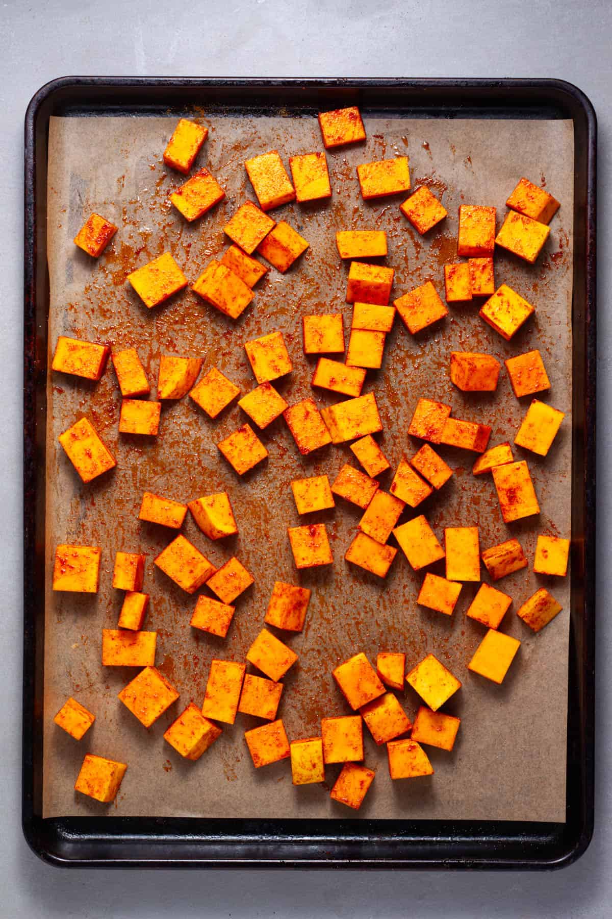 Cubes of butternut squash on a parchment paper lined baking sheet seasoned with spices.