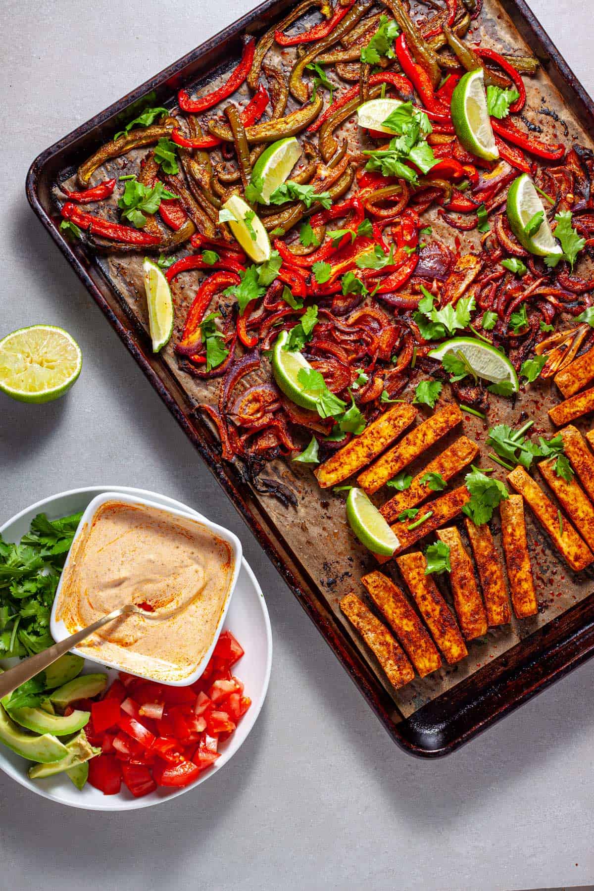 A sheet pan of tofu fajitas topped with cilantro and limes and a plate to the side with tomatoes, cilantro and a sour cream sauce.