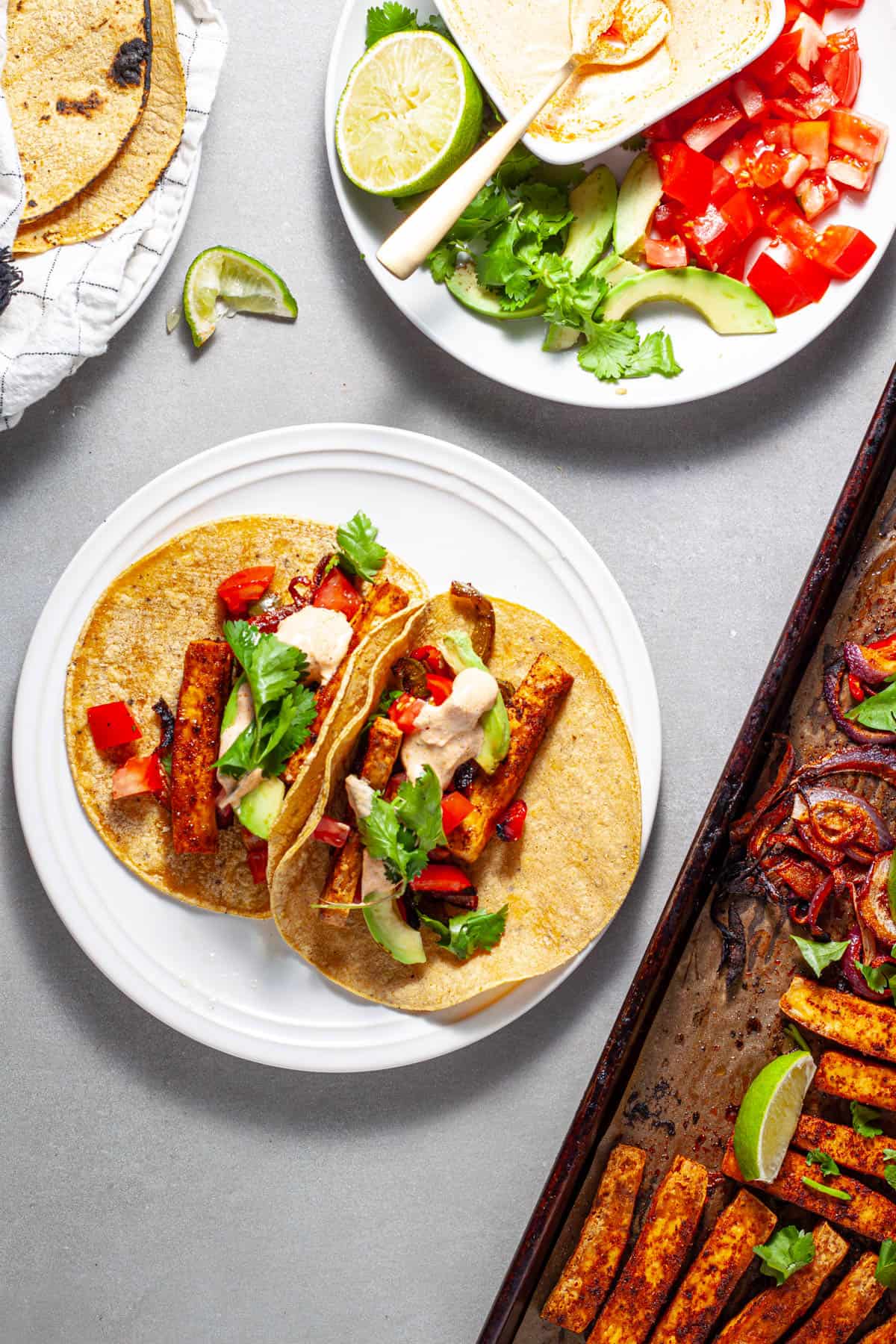 Sheet pan tofu fajita on a gray table with a small plate serving topped with cilantro, tomatoes and avocado.