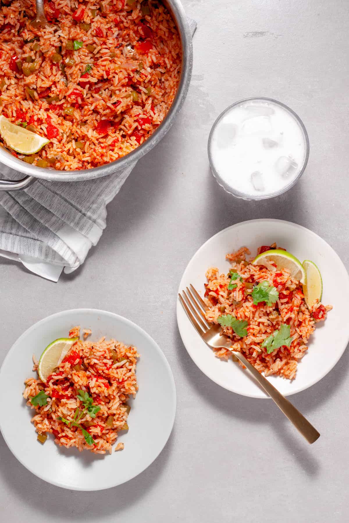 A couple plates of Tomato Rice topped with cilantro and limes and a saucepan of more rice to the side.
