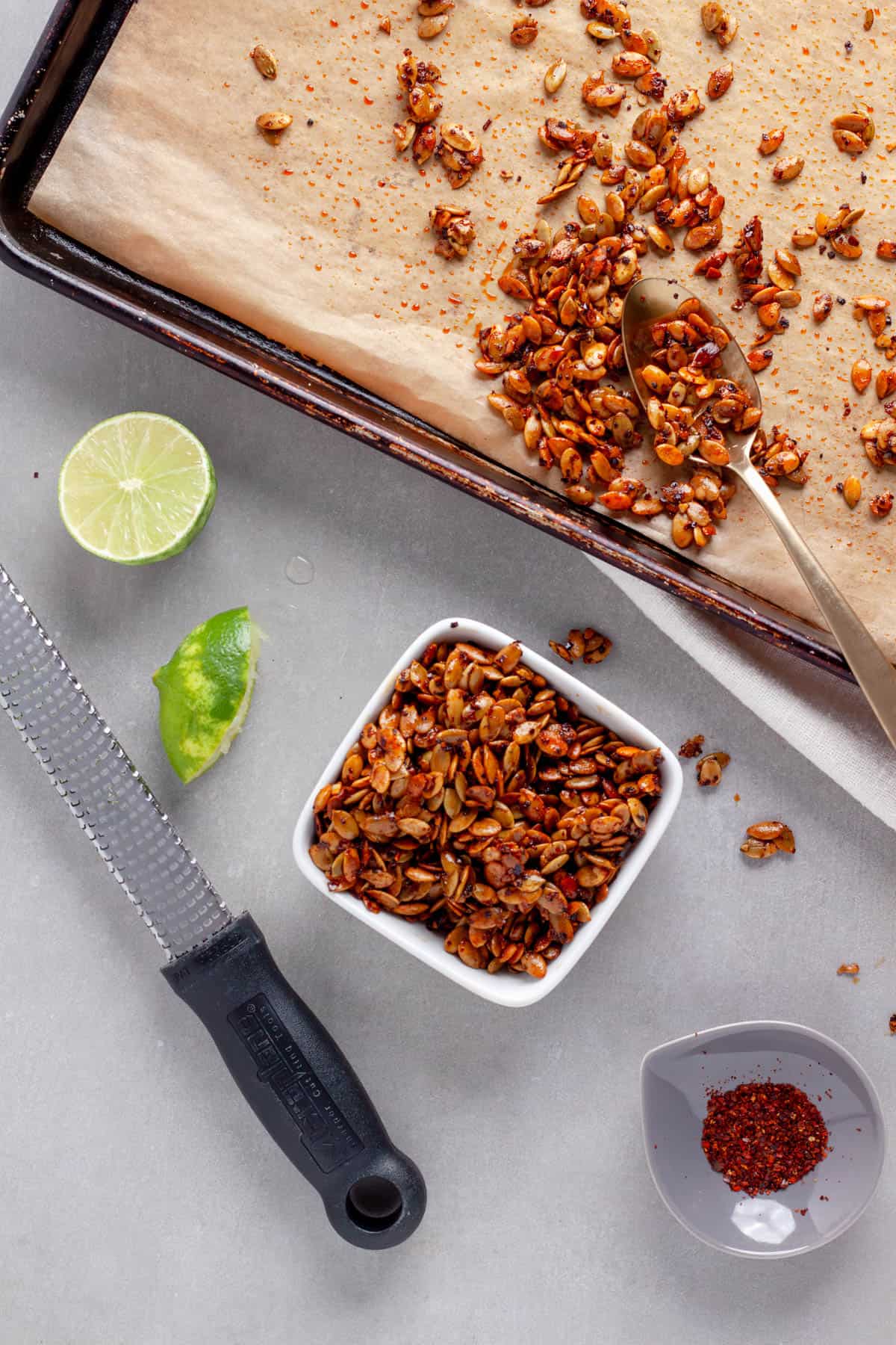 A small bowl of chili lime pepitas with a sheet tray of more to the side and a lime cut in half on the other side.