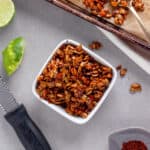 A small bowl of chili lime pepitas with a sheet tray of more to the side and a lime cut in half on the other side.