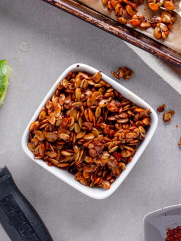 A small bowl of chili lime pepitas with a sheet tray of more to the side and a lime cut in half on the other side.
