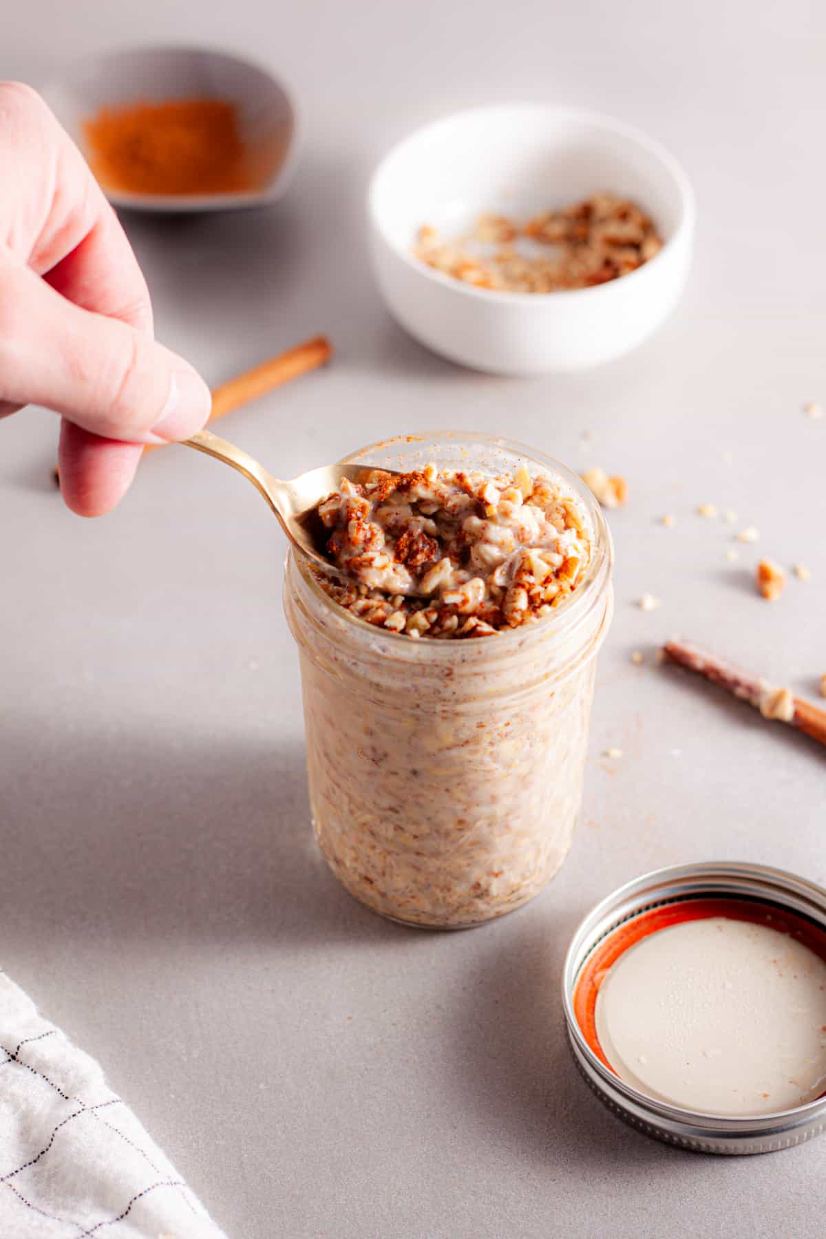 A glass mason jar with cinnamon overnight oats and a hand taking a spoonful out to eat.