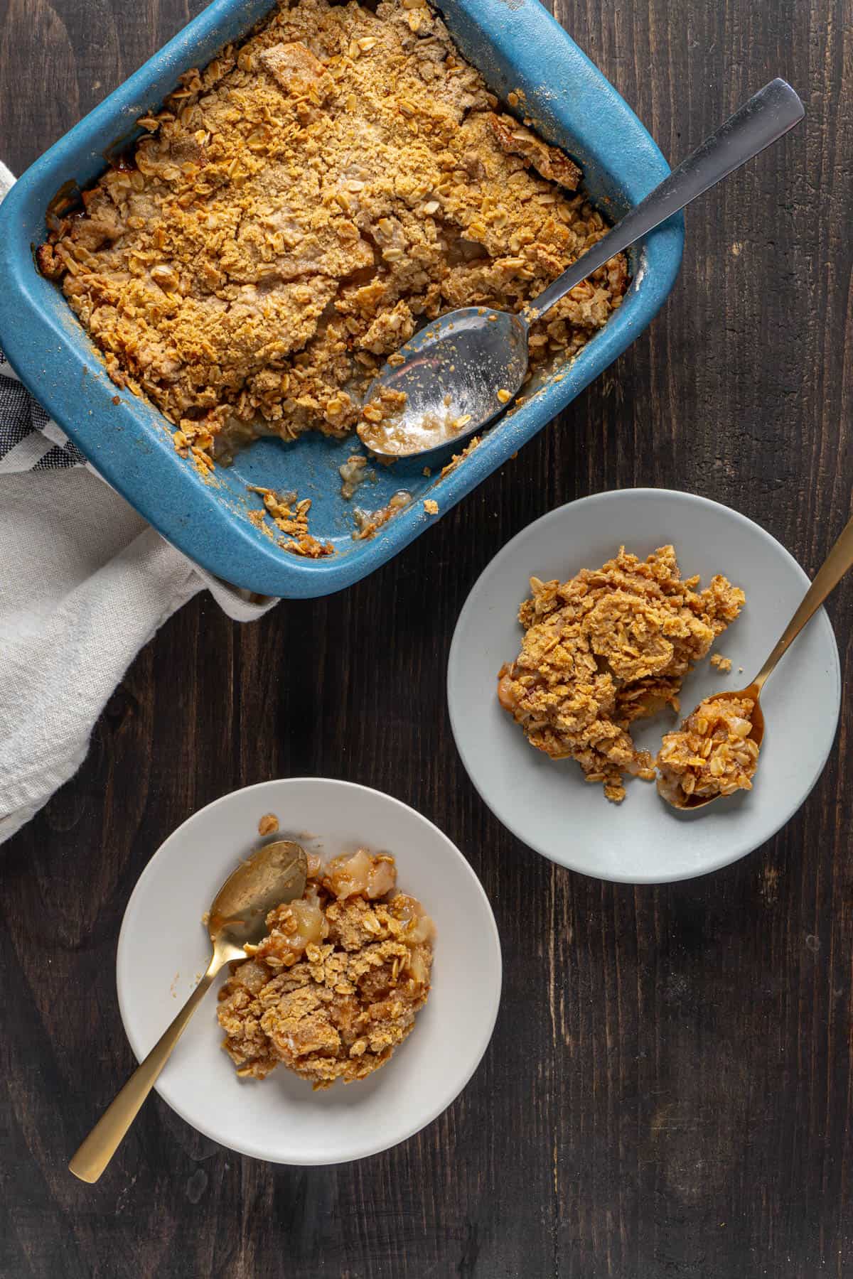 A couple portions of gluten-free apple crisp on plates with a baking dish to the background.