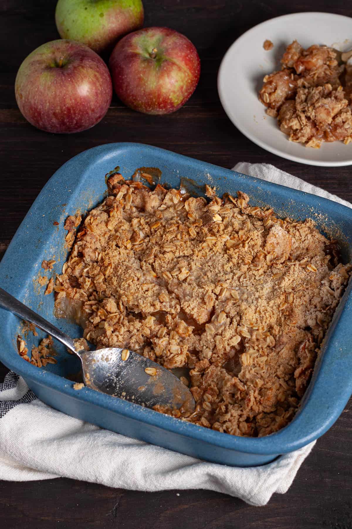 Gluten-free apple crisp in a blue baking dish with a serving and apples in the background.