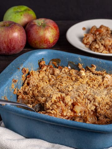 A blue baking dish with gluten-free apple crisp and apples in the background.
