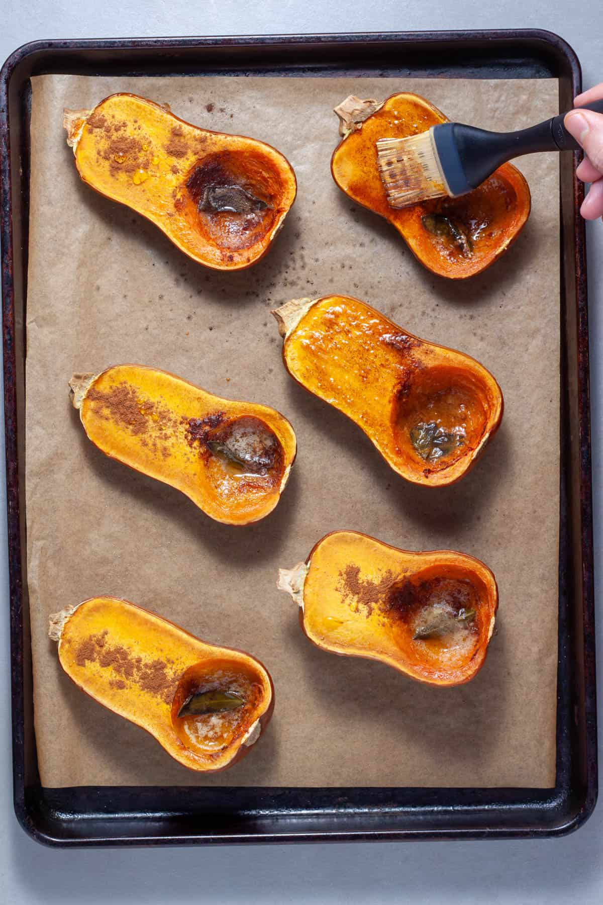 Roasted honeynut squash on a baking sheet getting brushed with melted butter and sage.