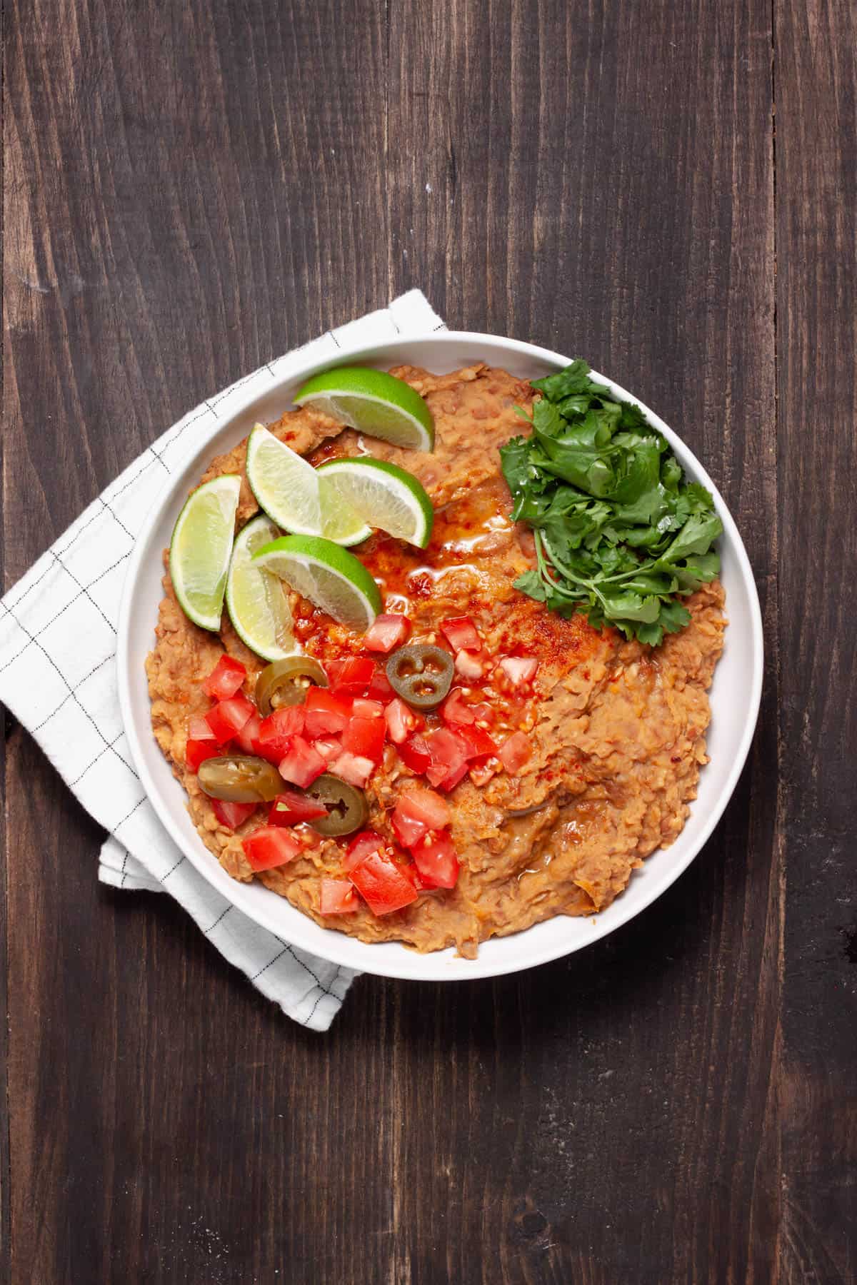 A bowl of vegan refried beans topped with cilantro, tomatoes, lime wedges and pickled jalapenos.