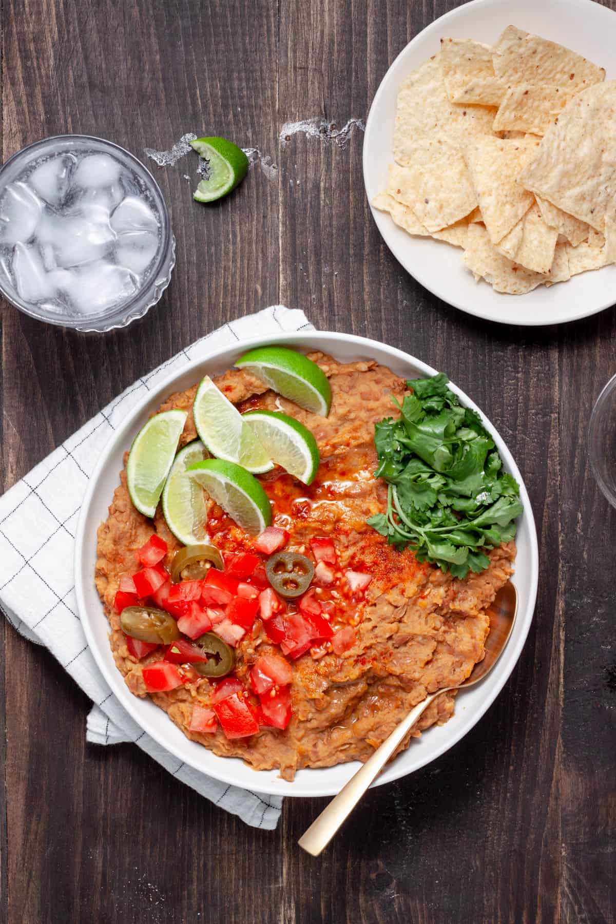 Vegan refried beans in a serving dish topped with lime wedges, cilantro leaves, chopped tomatoes and pickled jalapenos.