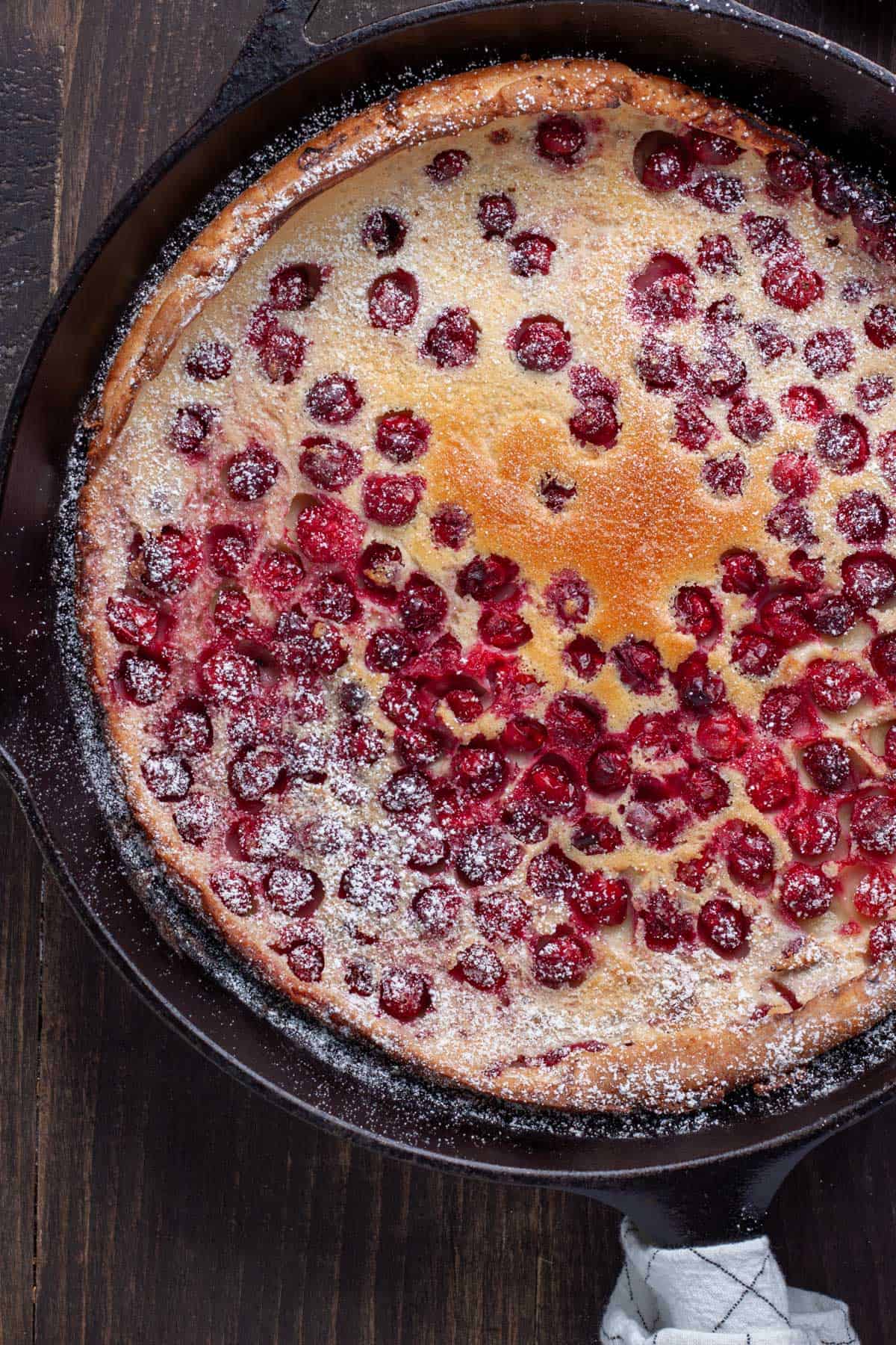 A cranberry Dutch baby in a cast iron skillet lightly dusted with powdered sugar.