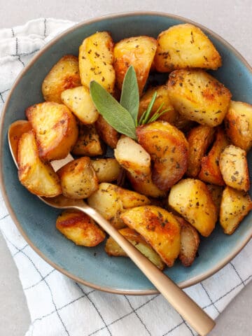 A green serving bowl of crispy herb roasted potatoes with a gold spoon.