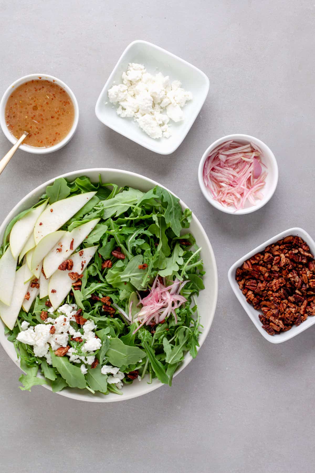 A big serving bowl with arugula pear salad with white balsamic dressing in a small bowl, along with spiced pecans, pickled shallots and goat cheese.