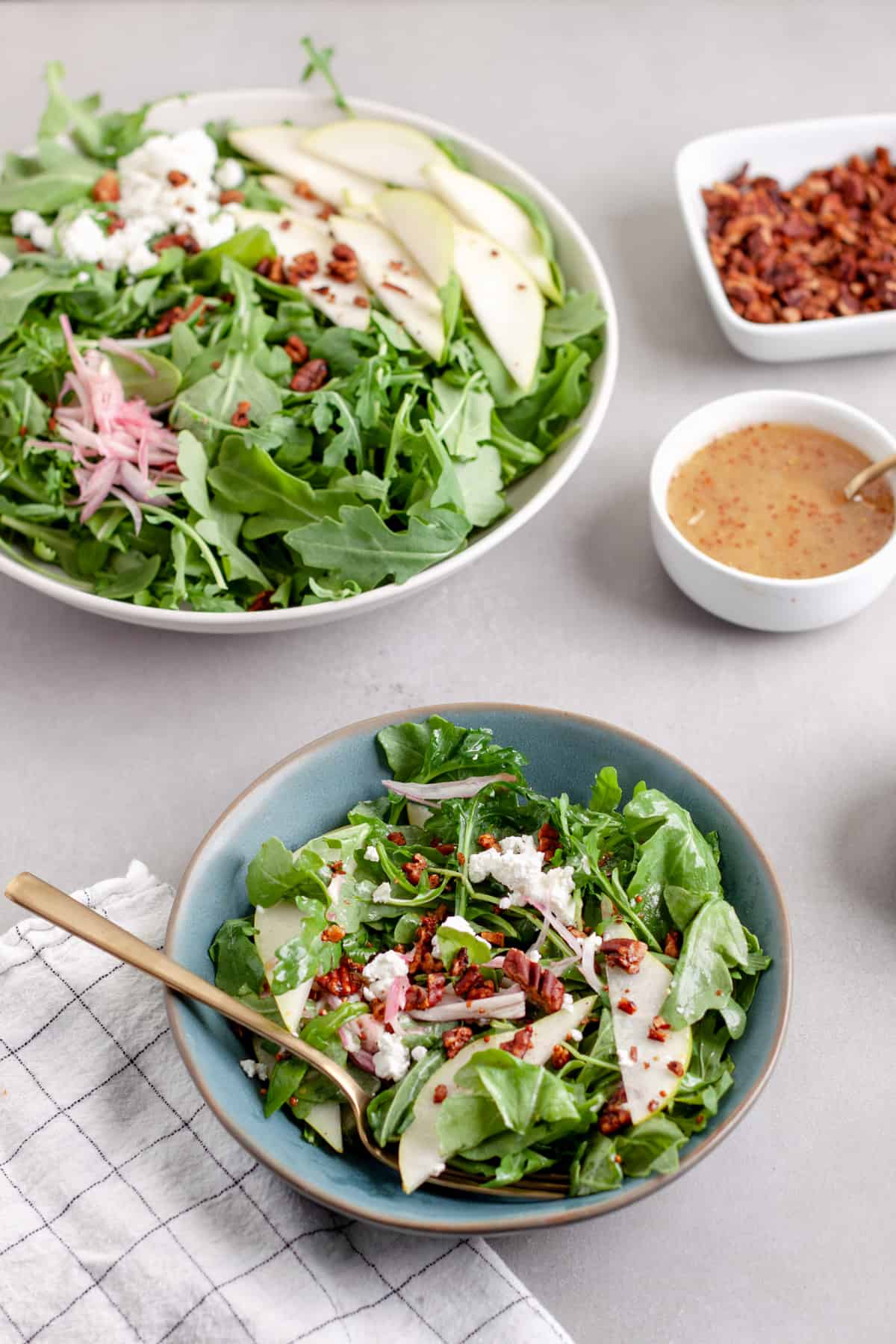 An arugula pear salad in a large bowl with a serving in a smaller blue bowl, topped with goat cheese and shallots.
