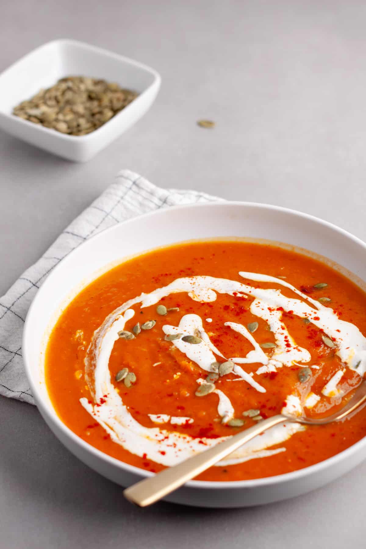 A bowl of butternut squash and red pepper soup garnished with sour cream, pepitas and paprika.