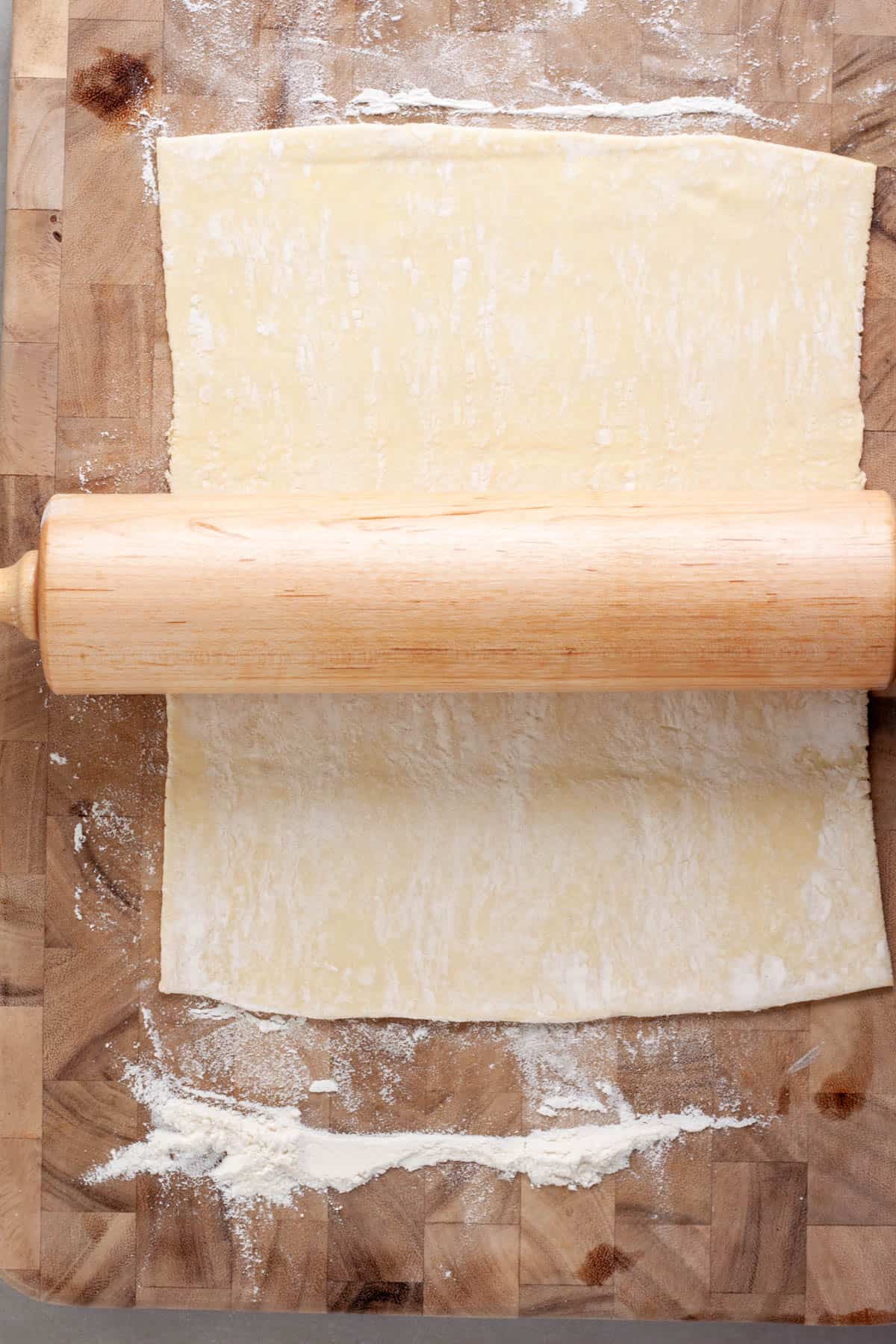 Puff pastry getting rolled on a wooden cutting board by a rolling pin.