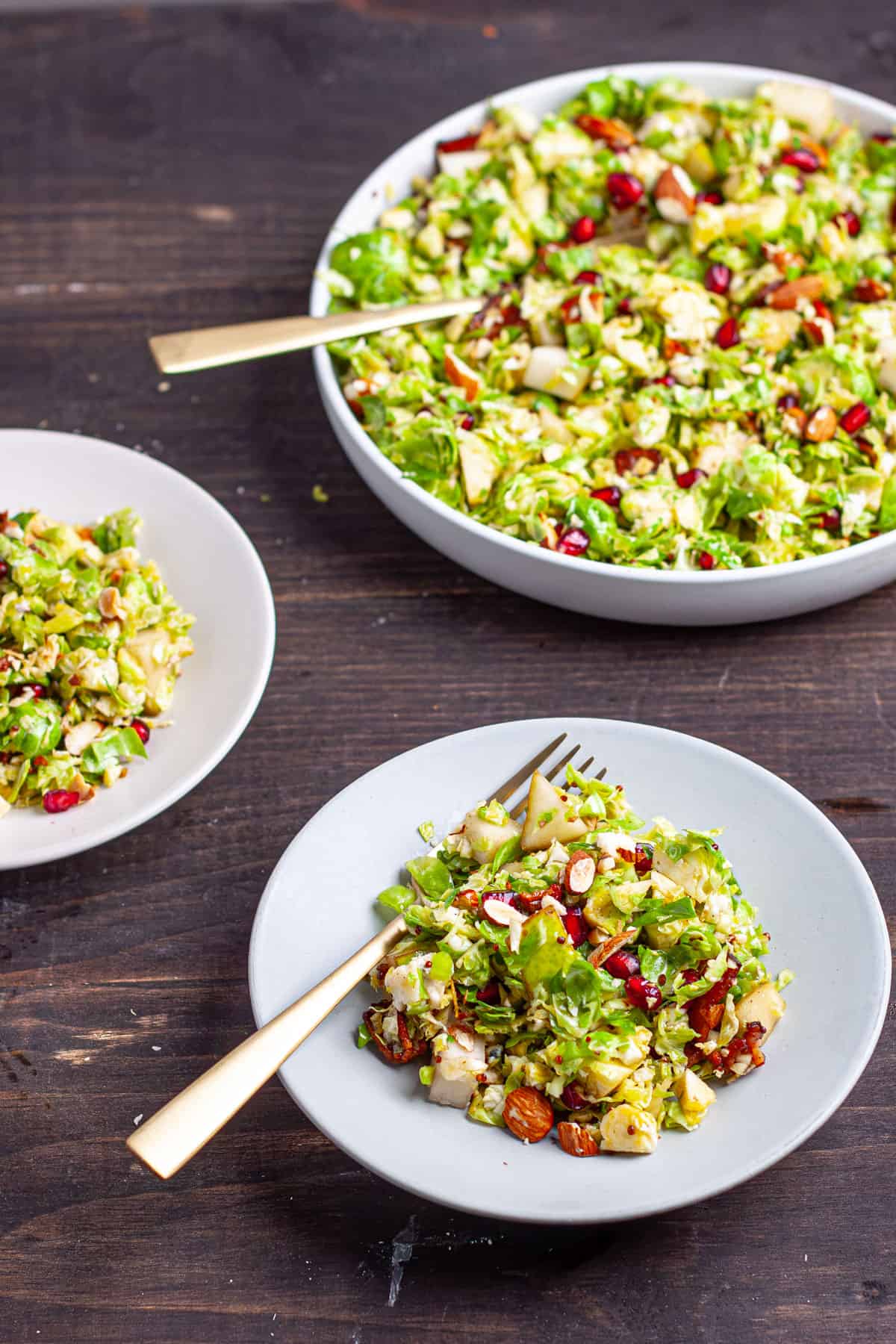 A large bowl of brussels sprouts salad with bacon and fig jam along with two servings on smaller plater on a dark table.