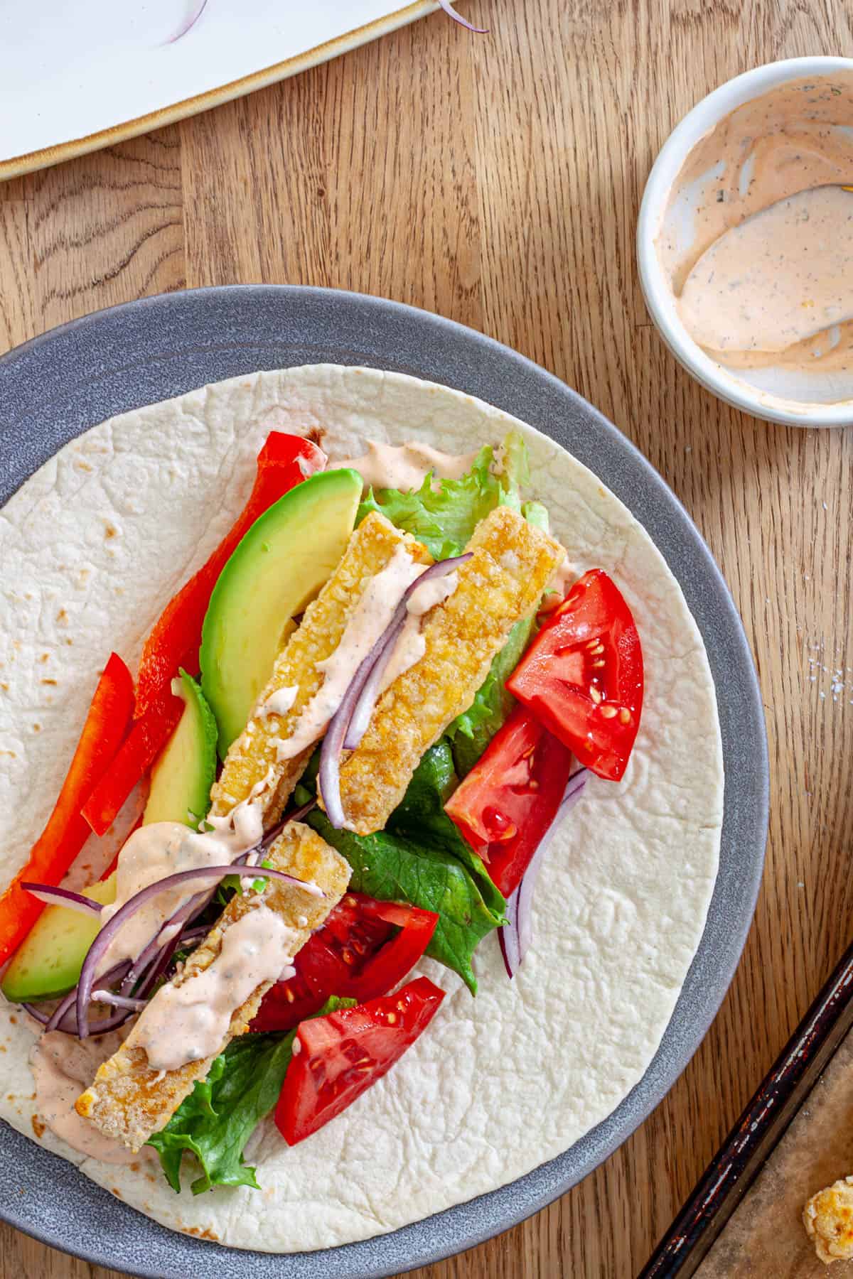 A plate with crispy buffalo tofu wrap topped with avocado, red peppers, lettuce, red onions and a homemade buffalo ranch sauce.