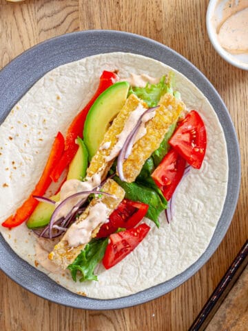 A plate with crispy buffalo tofu wrap topped with avocado, red peppers, lettuce, red onions and a homemade buffalo ranch sauce.
