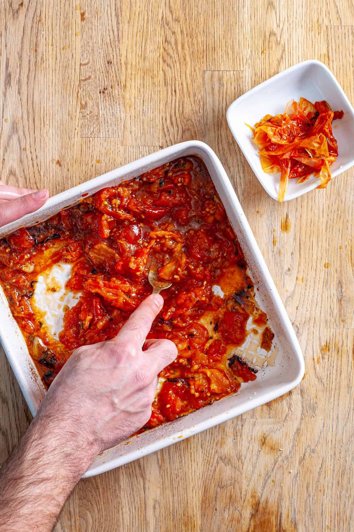A fork mashing roasted tomatoes and garlic in a white baking dish.