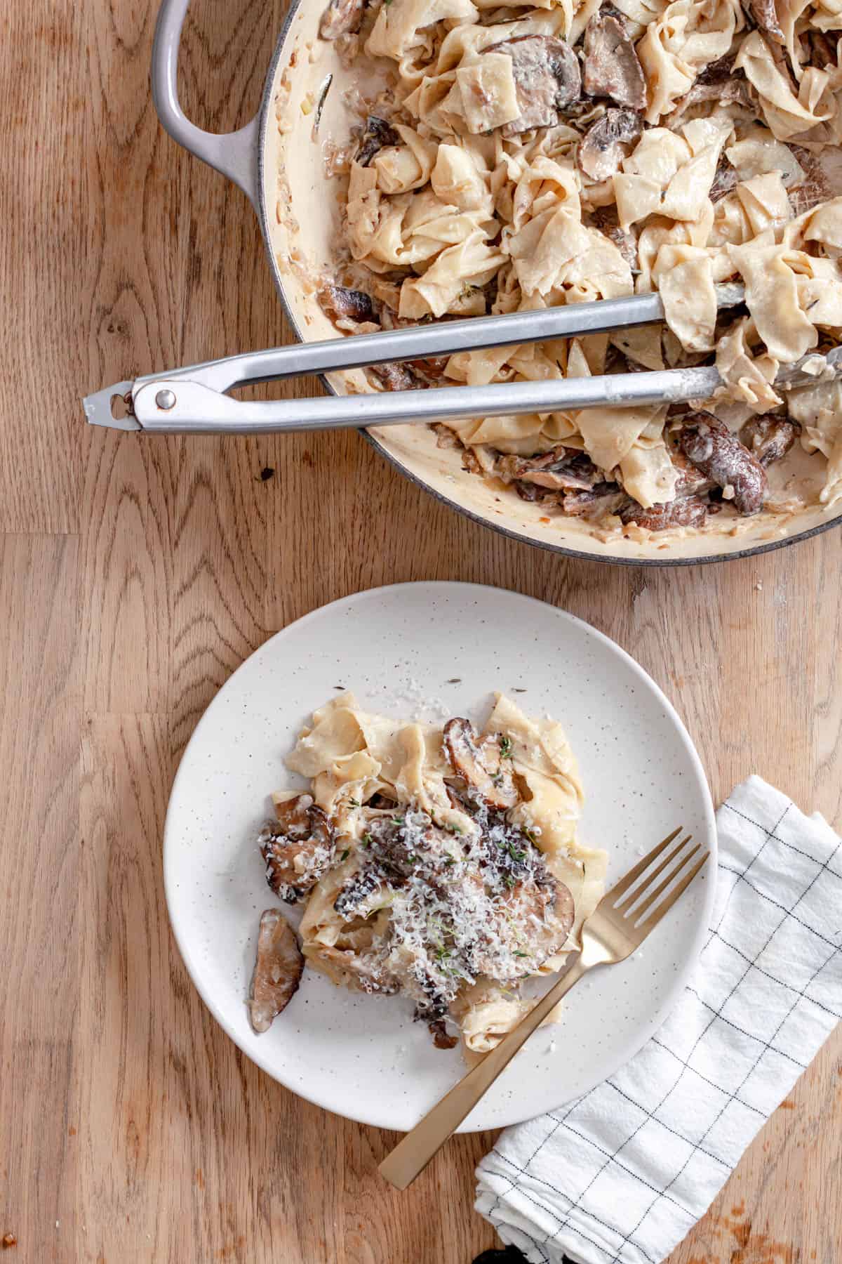 A serving of mushroom pappardelle on a white plate with a large skillet of more noodles to the side.
