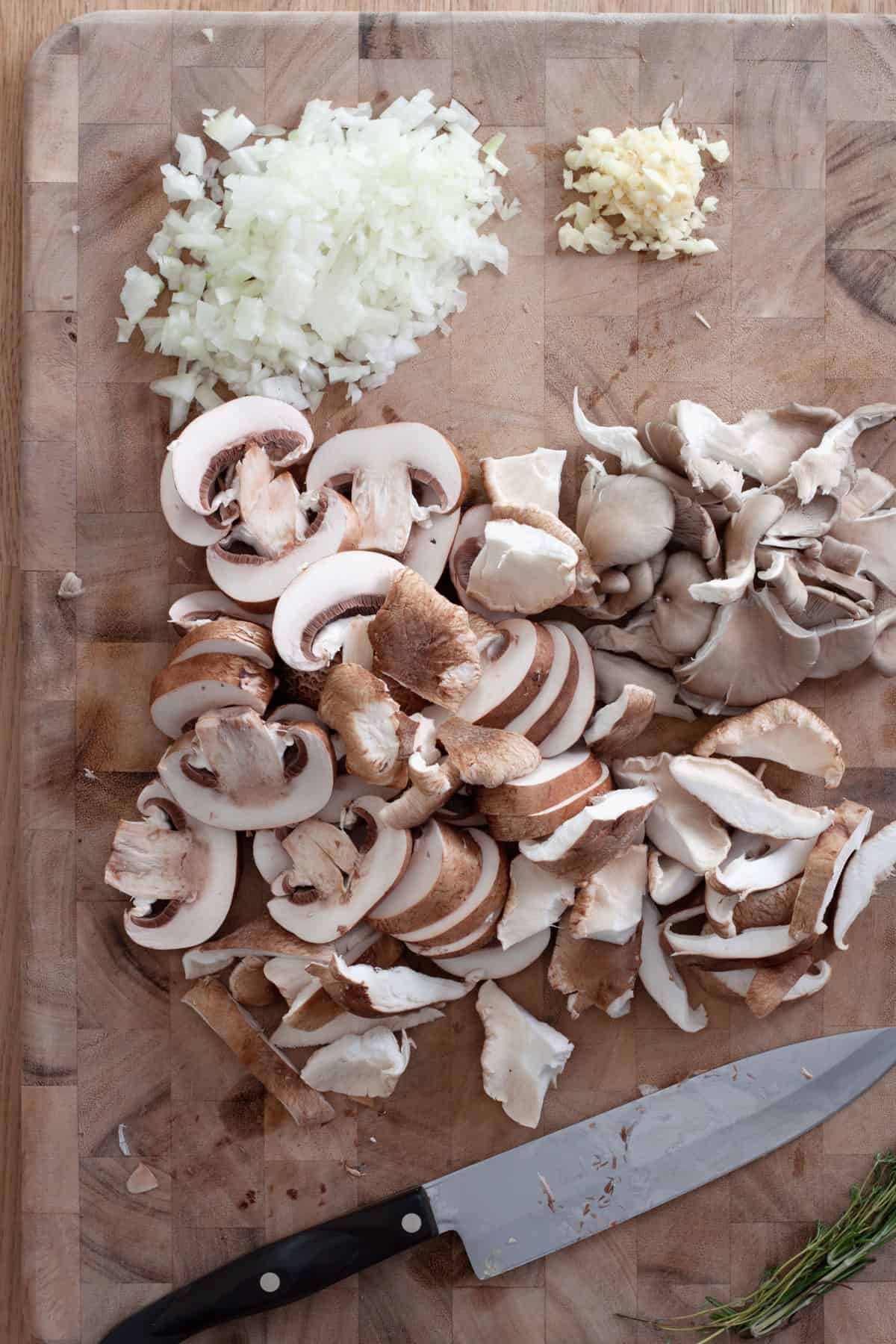 Various mushrooms sliced on a cutting board, along with minced garlic and a chopped onion.