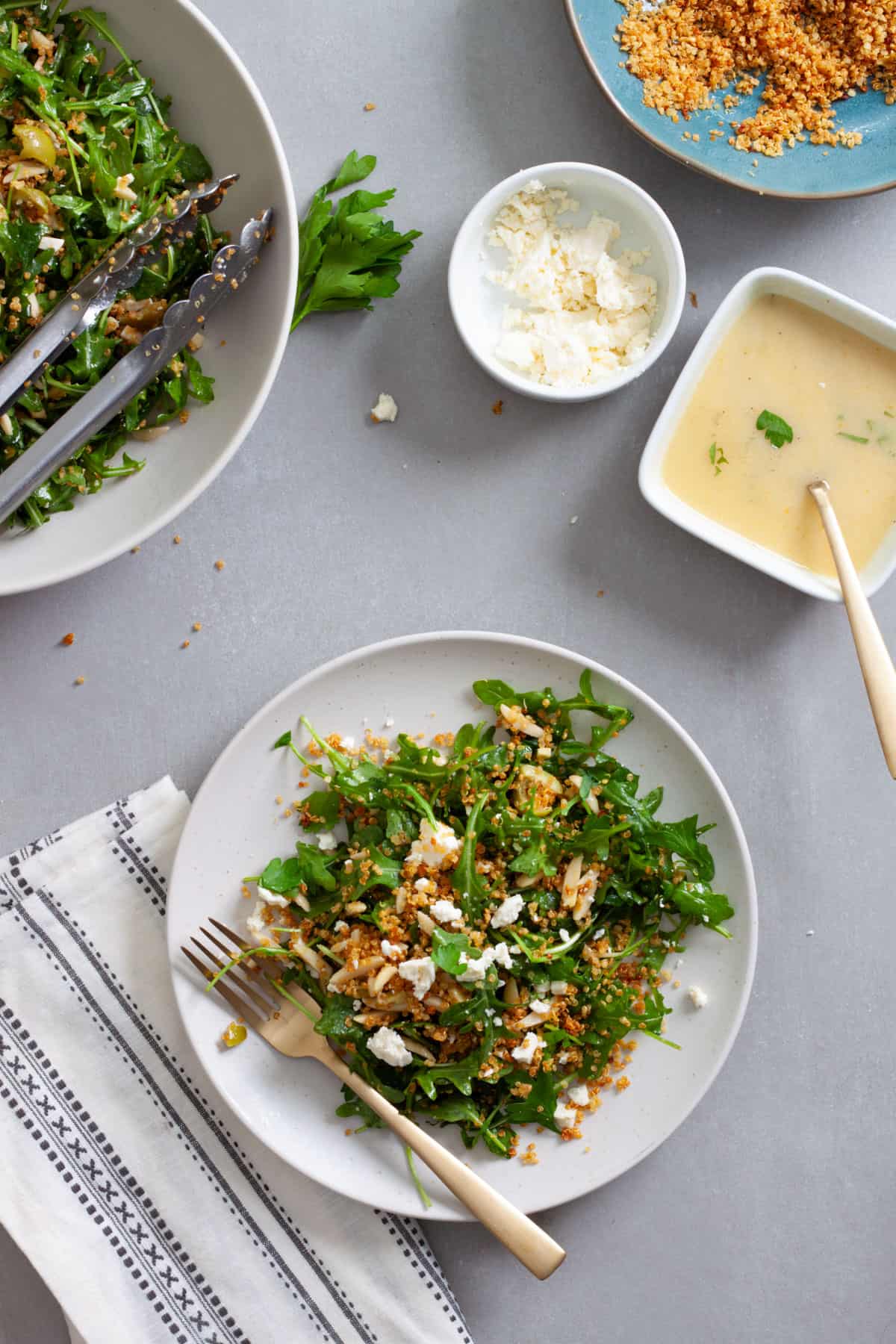 A serving of arugula and quinoa salad on a white plate with a gold fork on a gray table.