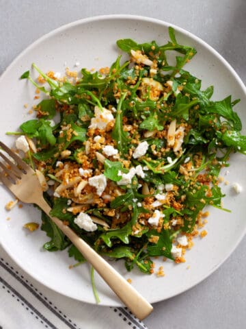 A serving of arugula and quinoa salad on a white plate with a gold fork on a gray table.