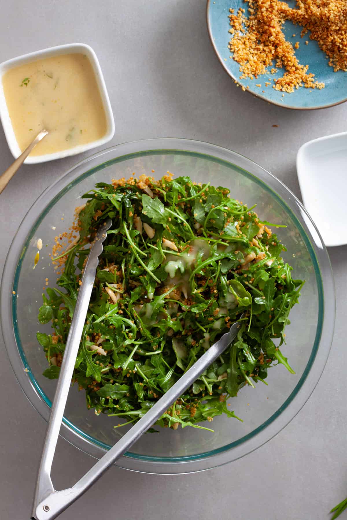Arugula and crispy quinoa in a large mixing bowl getting tossed by tongs.