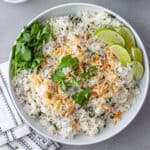 A white serving bowl with coconut cilantro rice topped with toasted shredded coconut and cilantro leaves with lime wedges to the side.