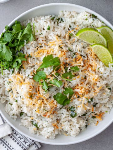 A white serving bowl with coconut cilantro rice topped with toasted shredded coconut and cilantro leaves with lime wedges to the side.