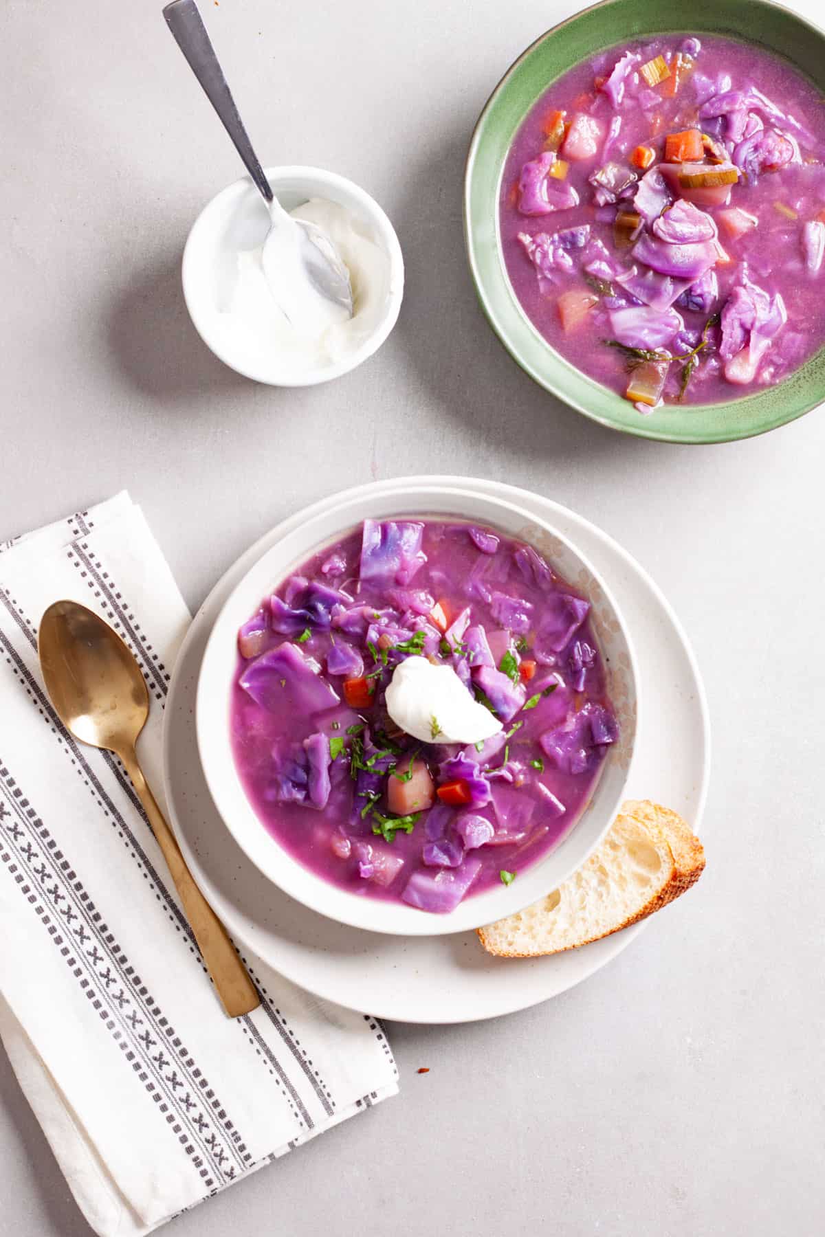 A couple servings of hearty red cabbage soup with one topped with sour cream and fresh herbs and a small bowl of sour cream to the side.