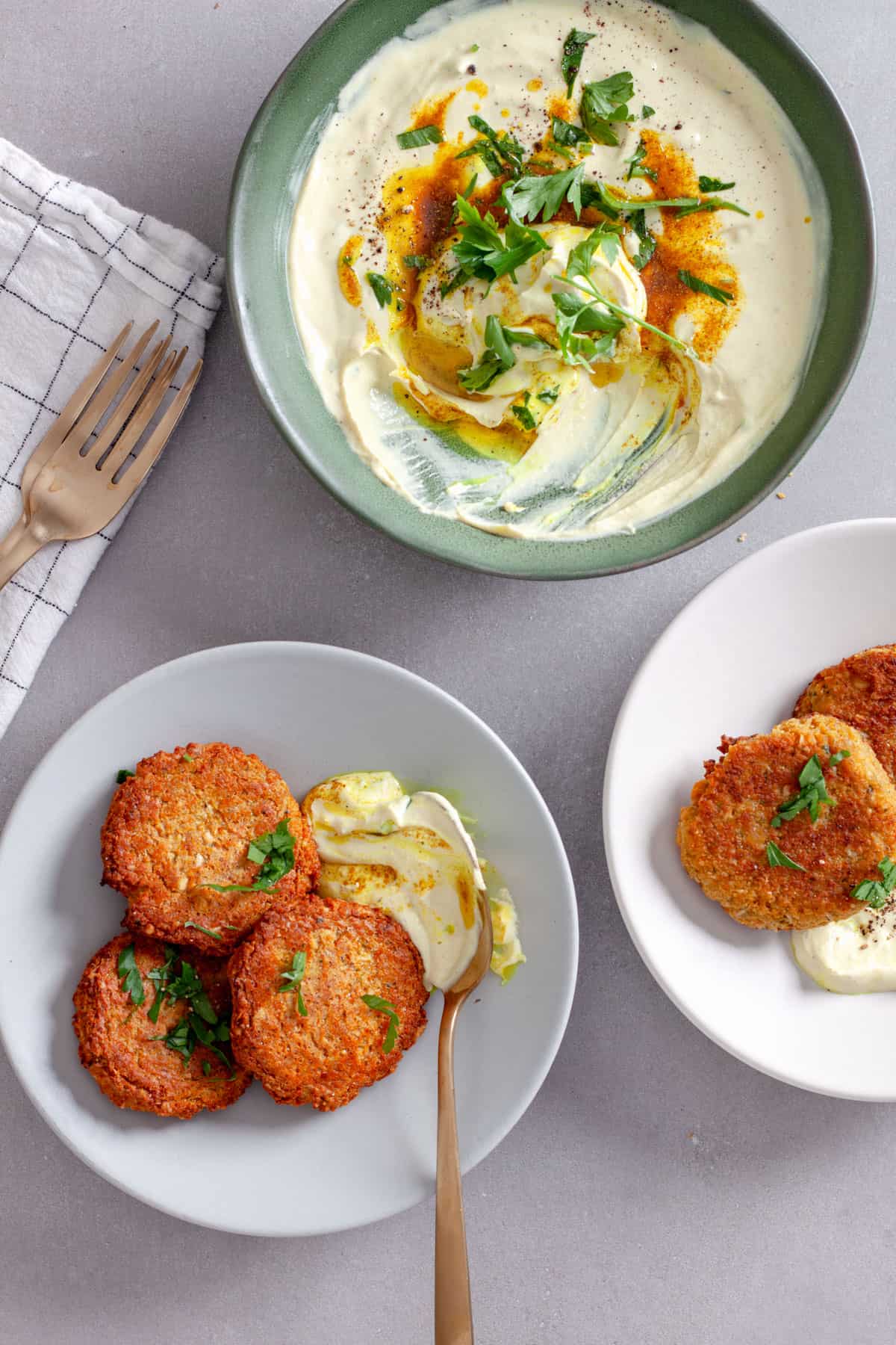 A couple plates of red lentil fritters topped with fresh herbs and a creamy tahini yogurt sauce on the side.