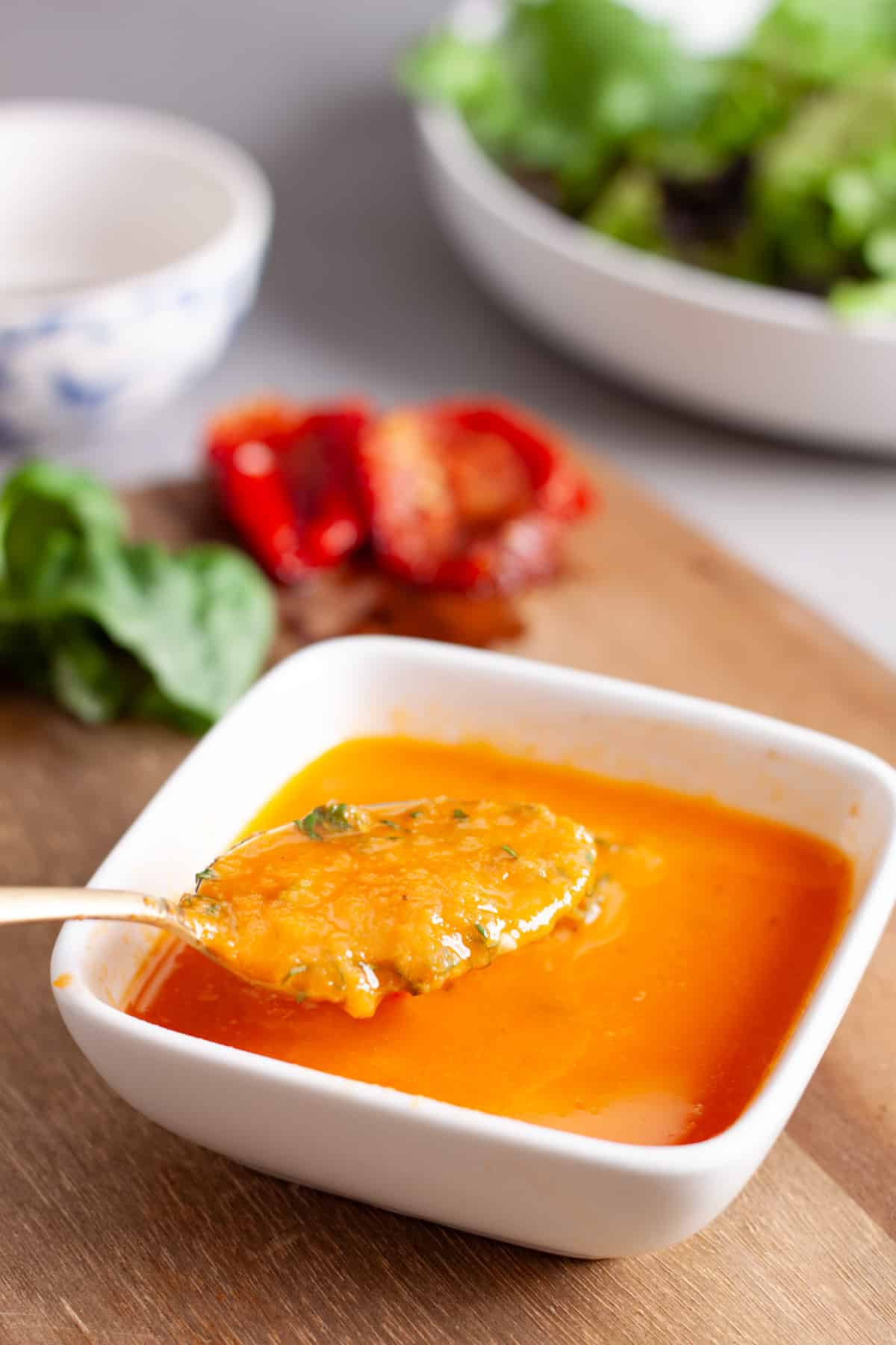 A small white bowl of sun-dried tomato vinaigrette with a spoon taking some out and a bowl of greens in the background.