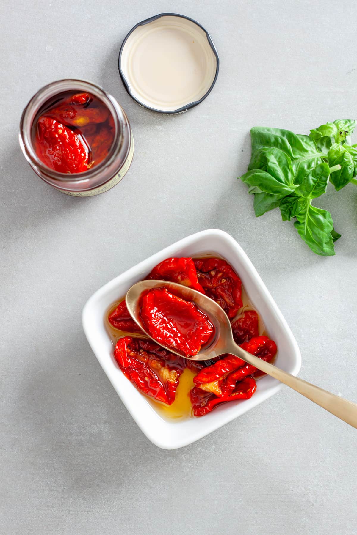 Whole sun-dried tomatoes in oil in a small white bowl with some basil off to the side.
