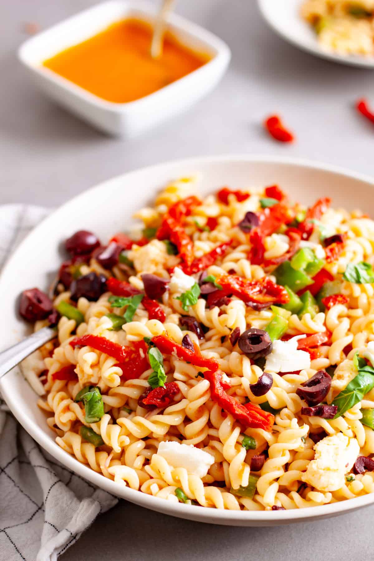 A large white bowl of sun-dried tomato pasta salad topped with olives, basil and cheese.