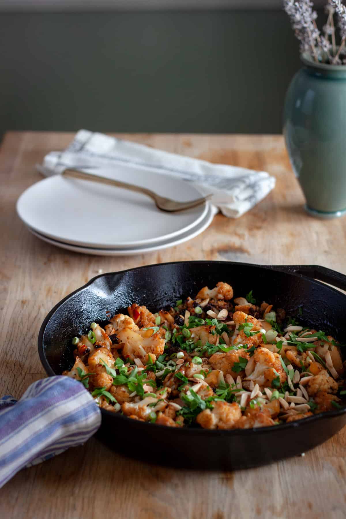 A cast iron skillet with sweet chili roasted cauliflower topped with parsley, almonds and raisins on a butcherblock countertop.
