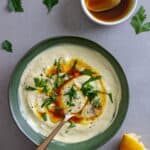 A bowl of tahini yogurt sauce topped with chopped parsley and cumin oil.