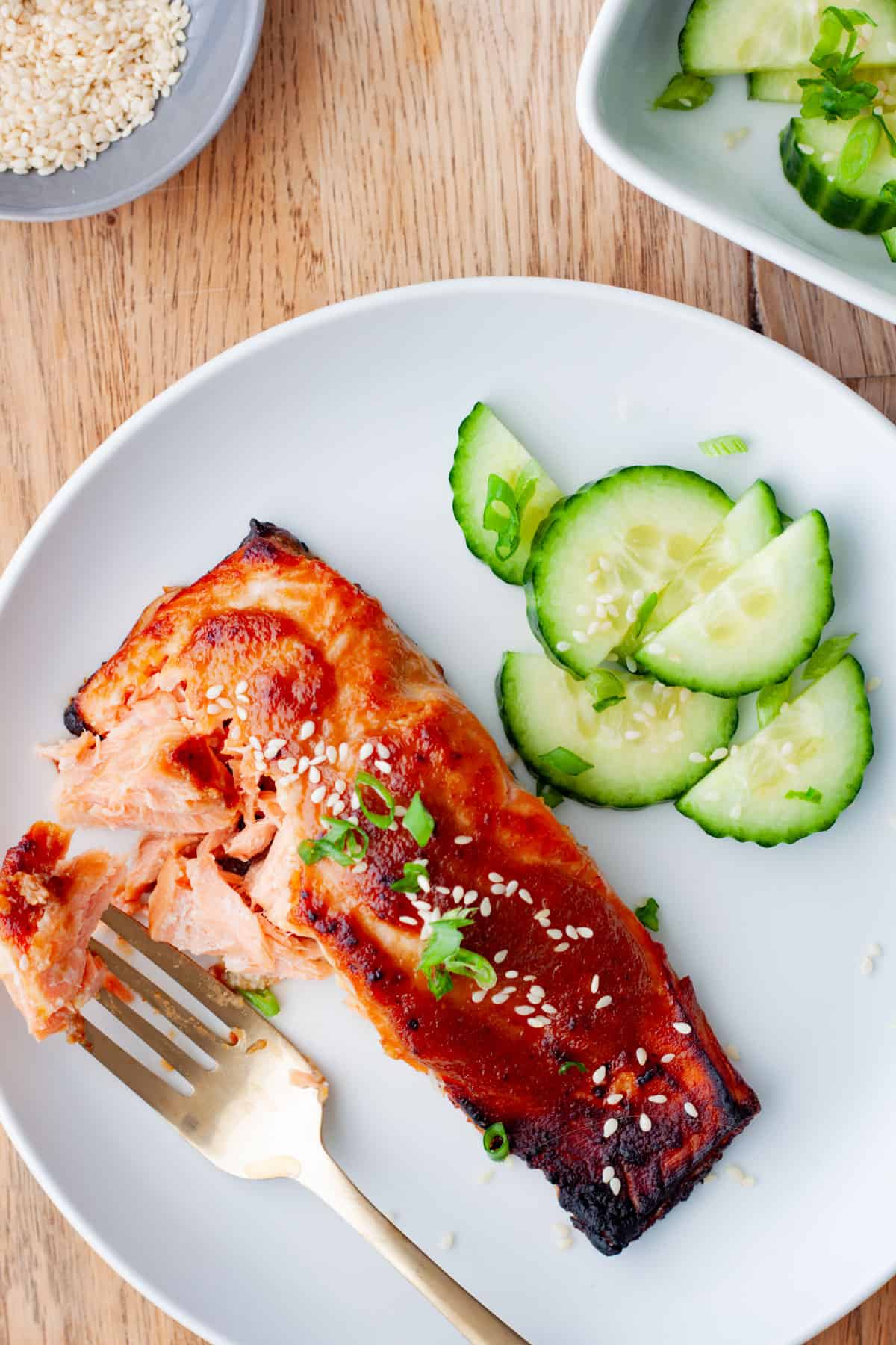 A fork taking a bite out of miso salmon with sliced cucumbers to the side.
