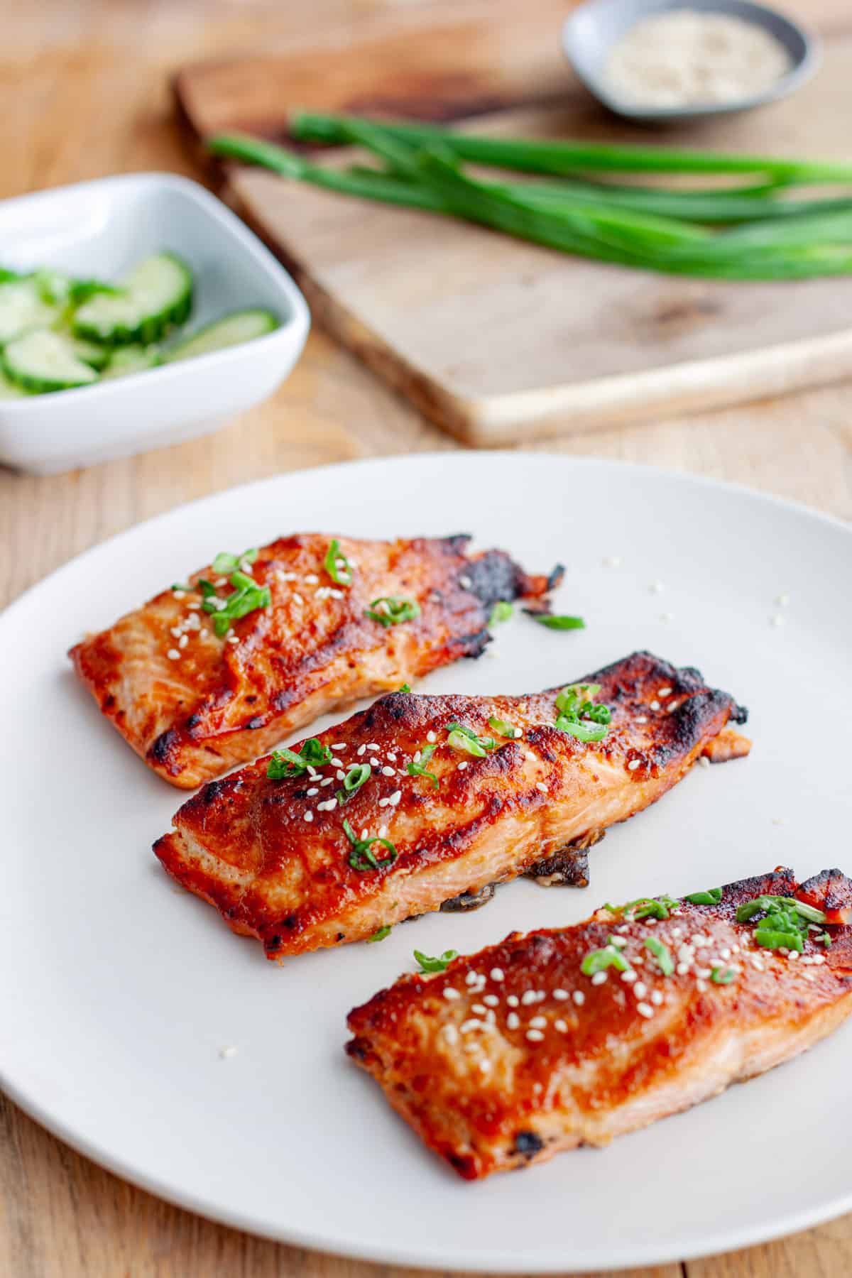 A platter of miso salmon on a butcherblock countertop with scallions and cucumbers in the background.