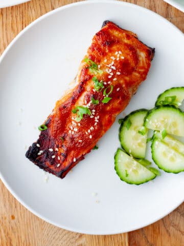 A white plate with a serving of miso salmon topped with sesame seeds and scallions and sliced cucumbers to the side.