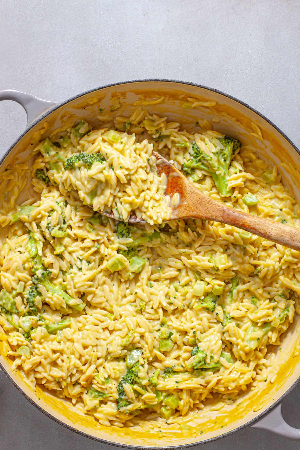A wooden spoon pulling out a serving of creamy broccoli orzo with cheddar cheese.