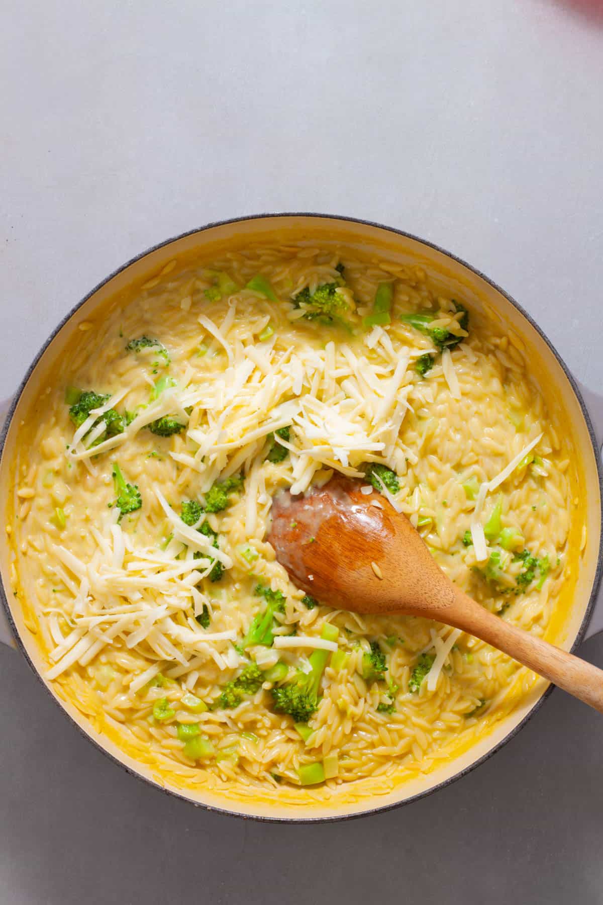 Shredded cheddar cheese getting stirred into a large skillet with broccoli orzo.