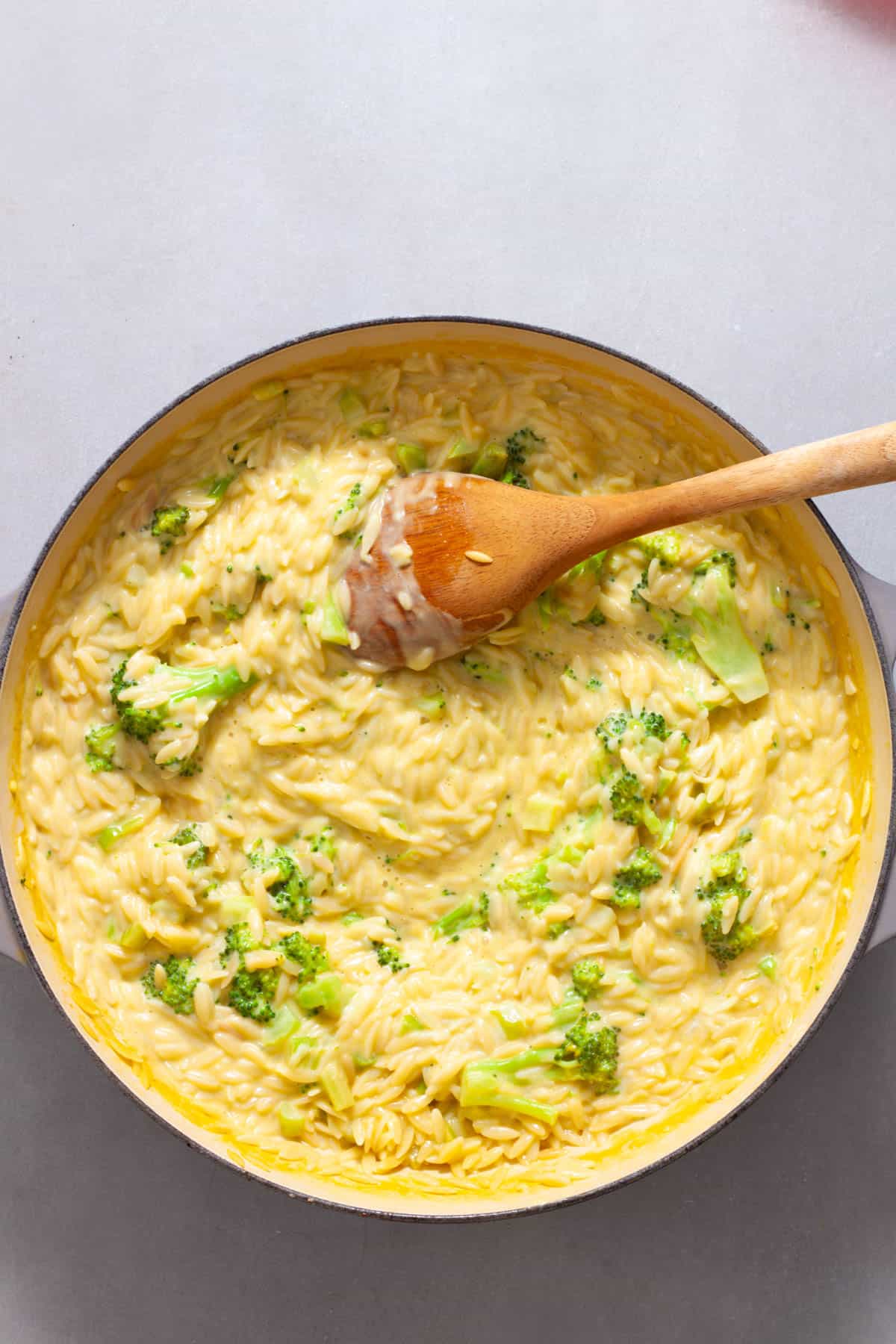 A wooden spoon finishing a large skillet of creamy broccoli orzo.