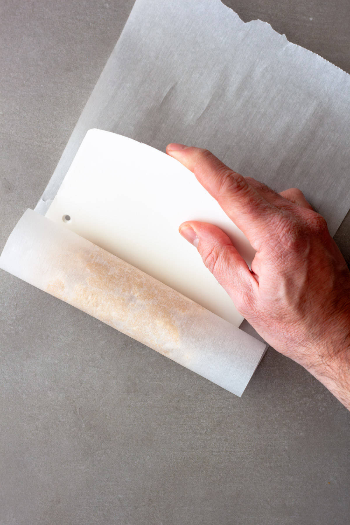 A hand pushing a bench scraper against a roll of miso butter in parchment paper.