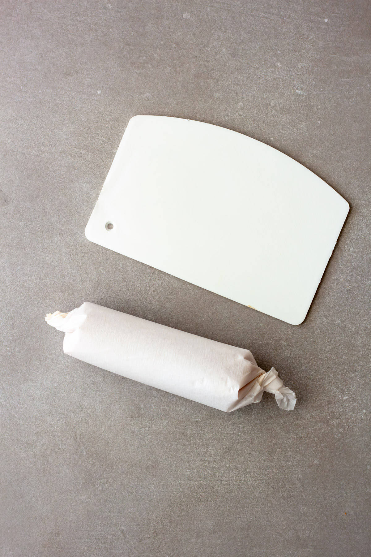 A roll of miso butter in parchment paper and a white bench scraper on a gray table.
