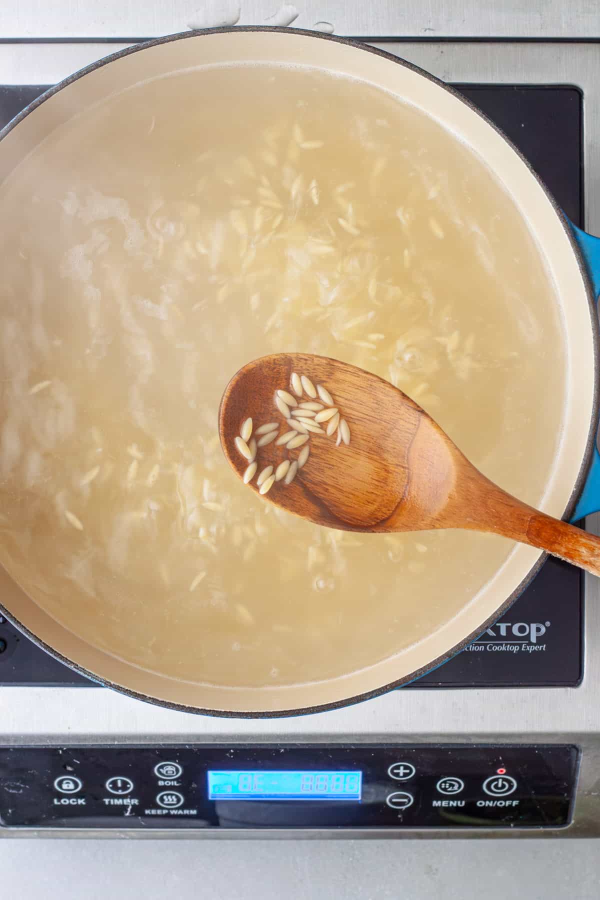 Orzo cooking in a large pot of water with a wooden spoon taking out some of the orzo.